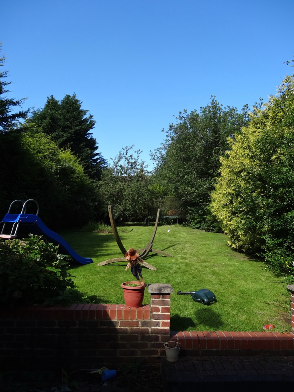 A tall view across the sunny garden towards the slide and hammock frame.