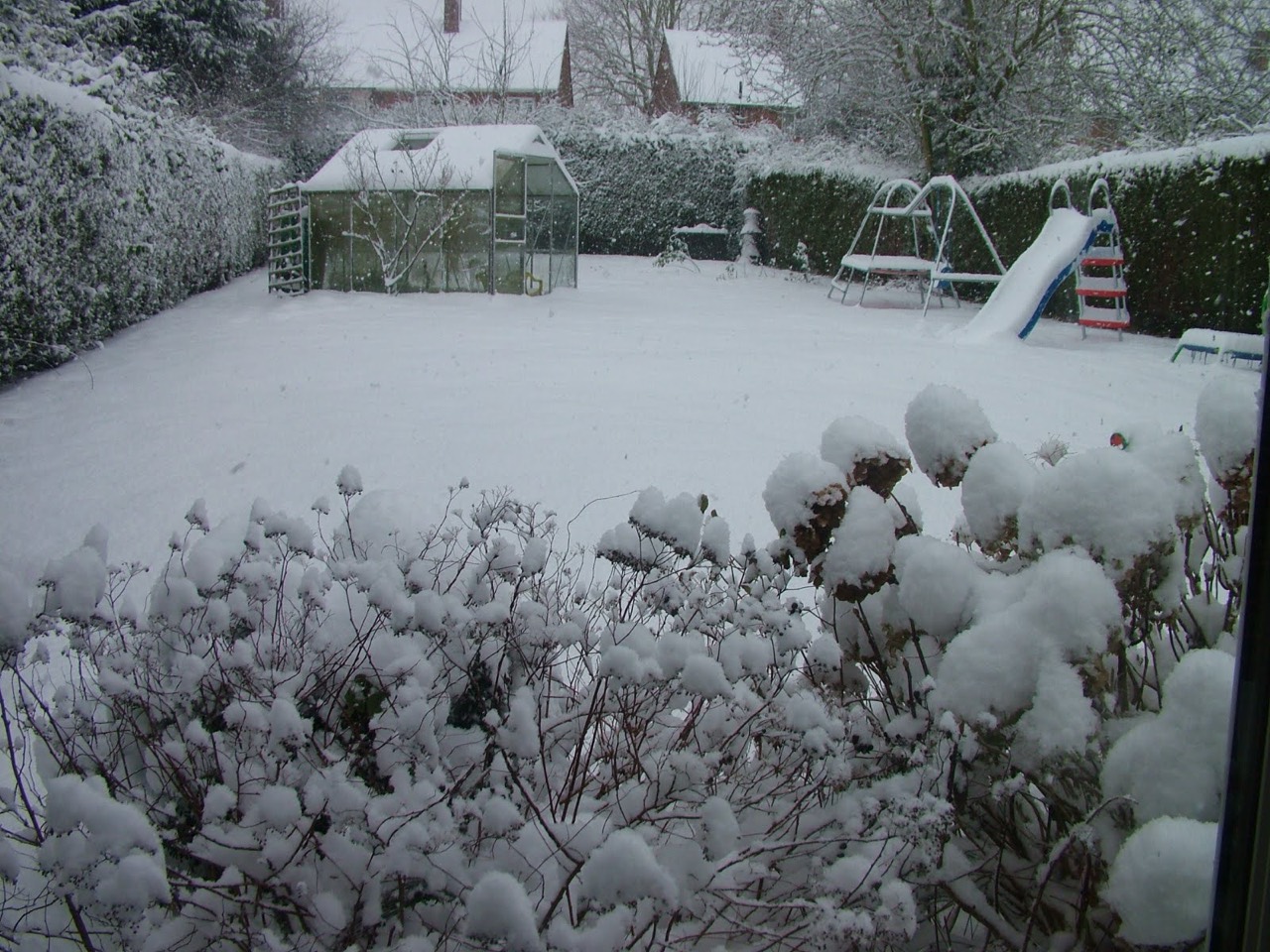 The childhood garden covered in snow, with the greenhouse, swings and slide in the distance.